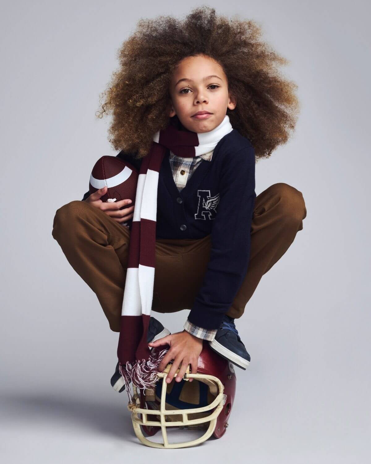 Child in sports-themed outfit with helmet and football on a plain background
