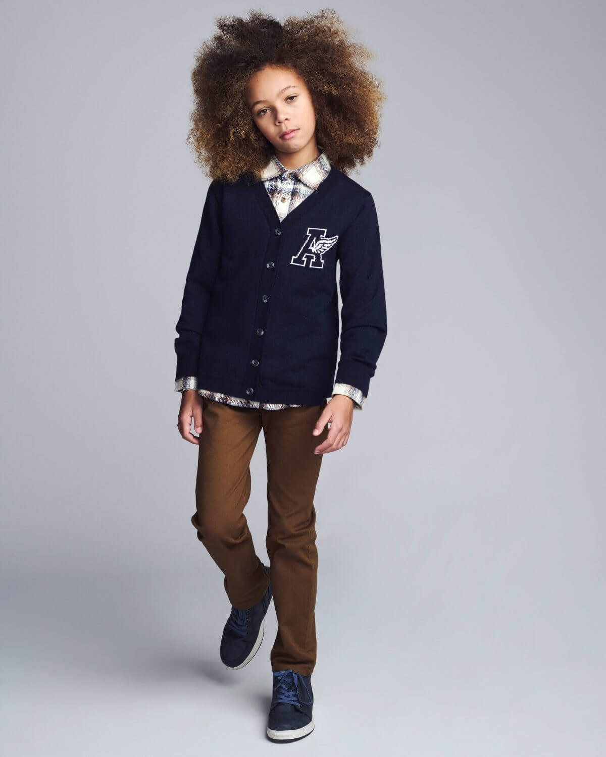 Child wearing a navy cardigan with a logo, plaid shirt, and brown pants on a gray background