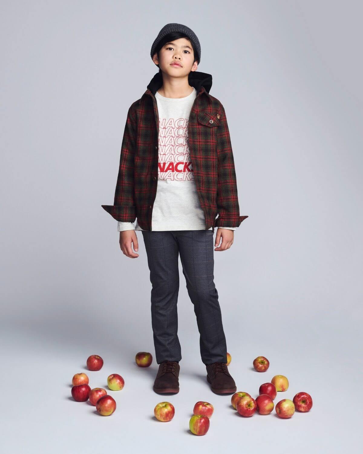 Child wearing a plaid shirt and jeans standing among apples on a gray background