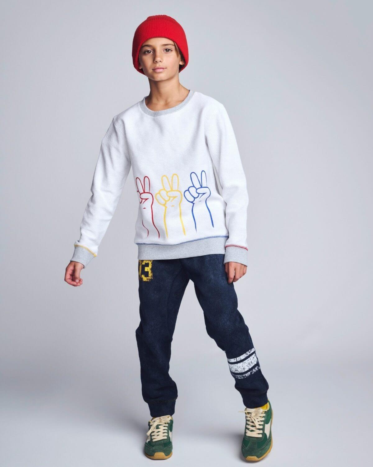 Person wearing a white sweatshirt with colorful designs, dark jeans, and green sneakers on a plain background