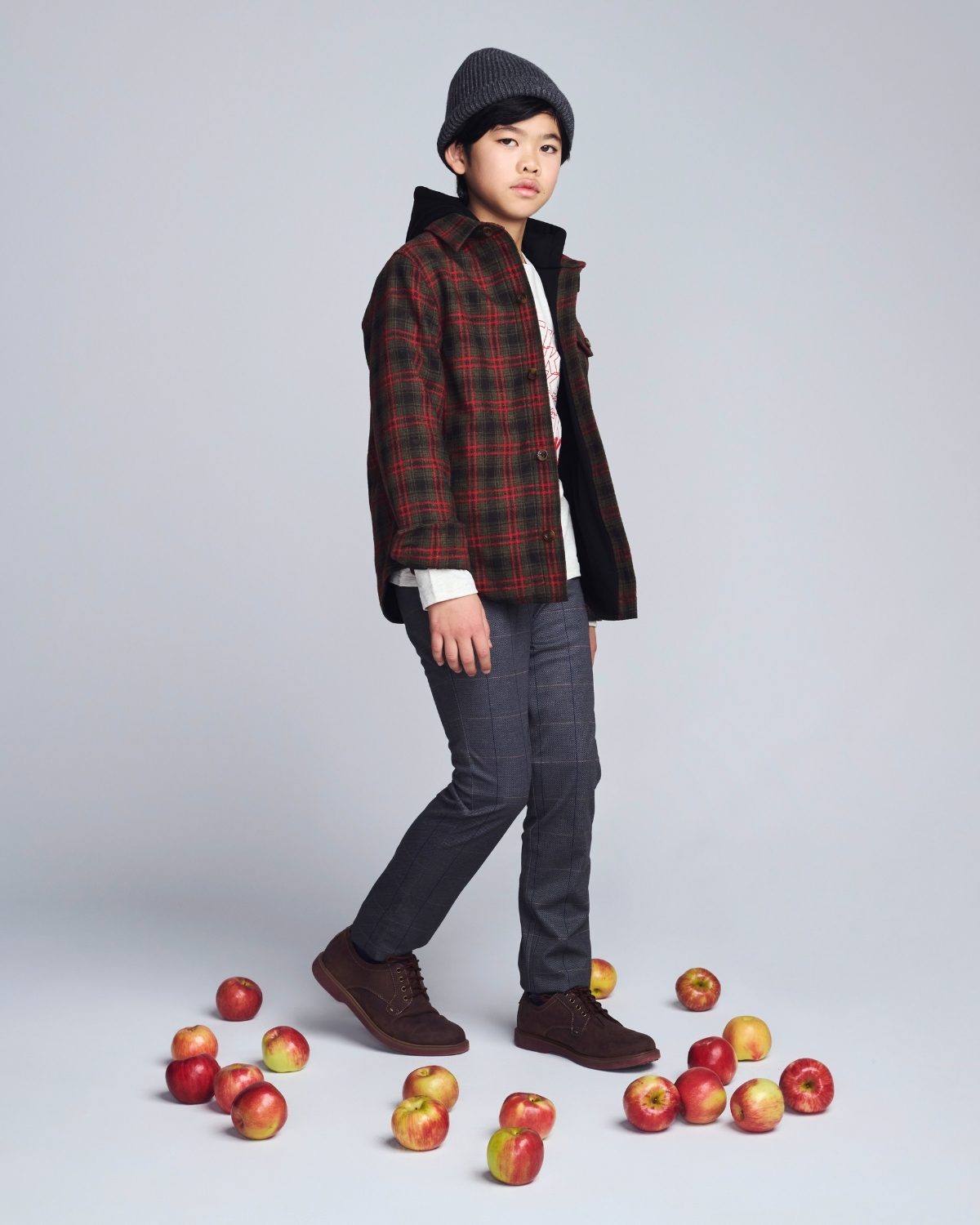 Person wearing a plaid jacket and dark pants standing among apples on a light gray background