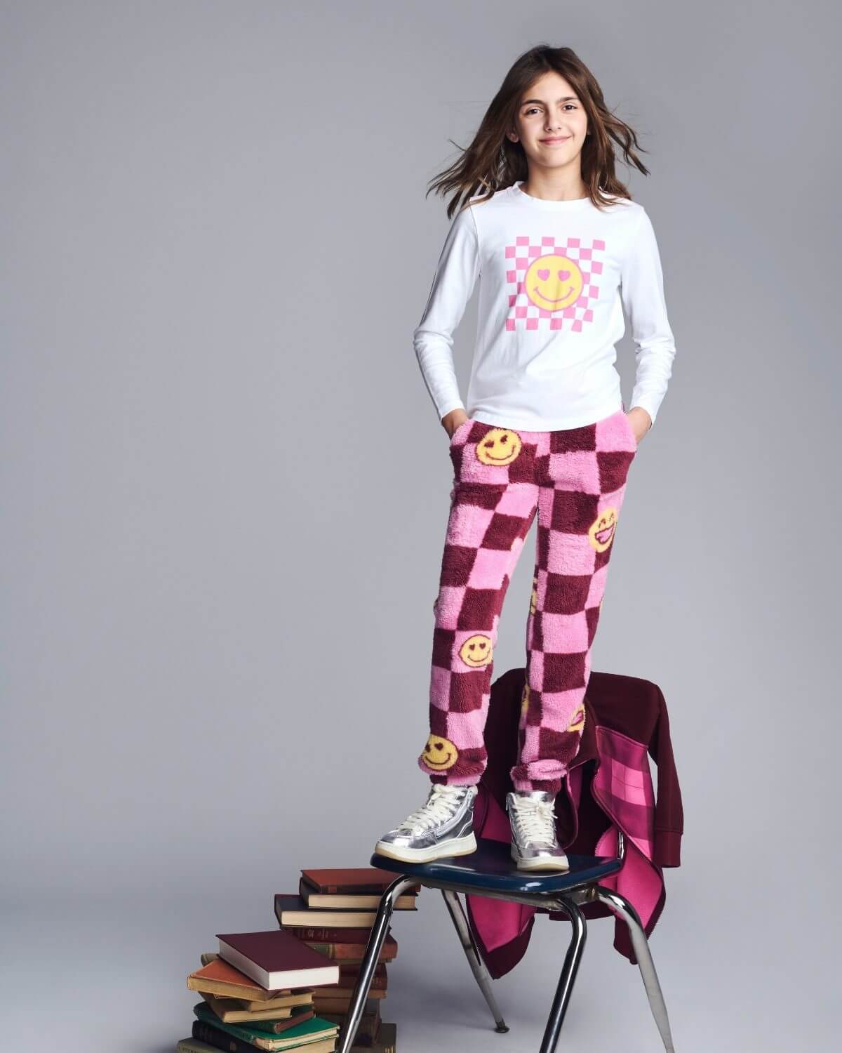 Person wearing a white shirt with smiley face design and pink checkered pants, standing on books.