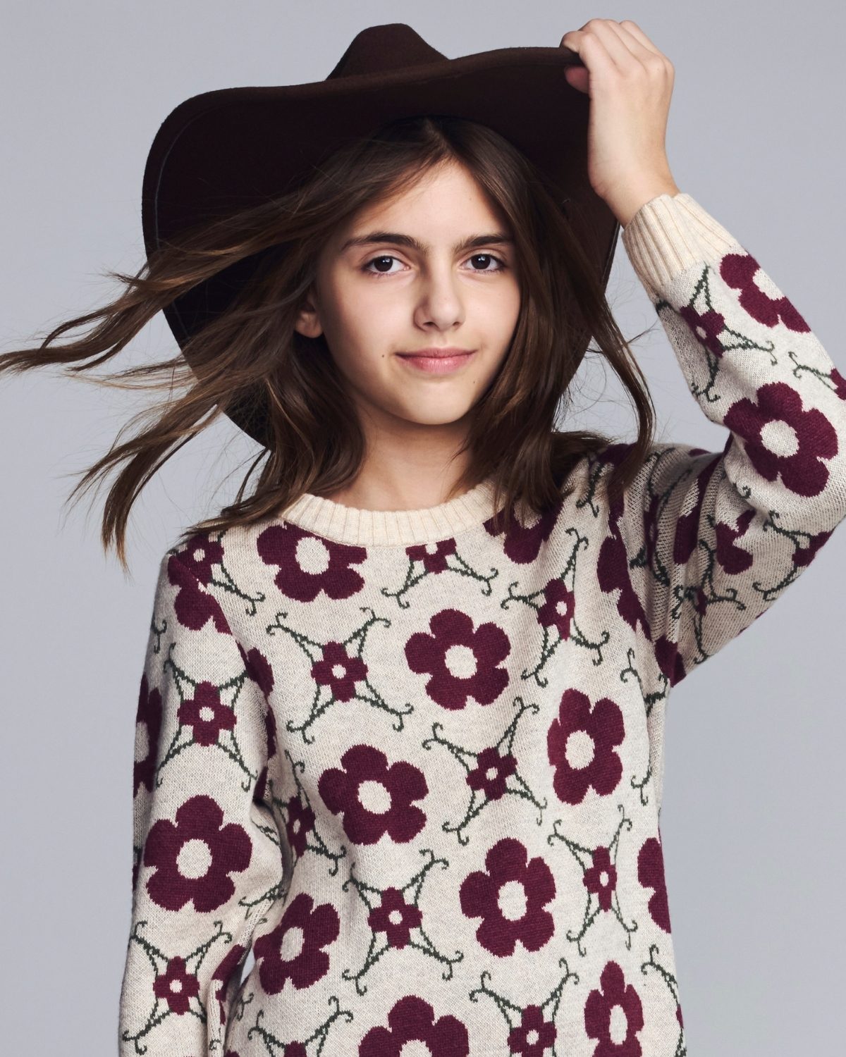 Young girl wearing a floral sweater and brown hat against a gray background