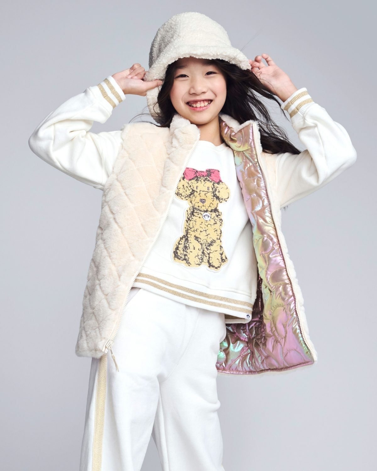 Person wearing a white coat with colorful lining, white pants, and a white shirt with a bear design on a gray background.