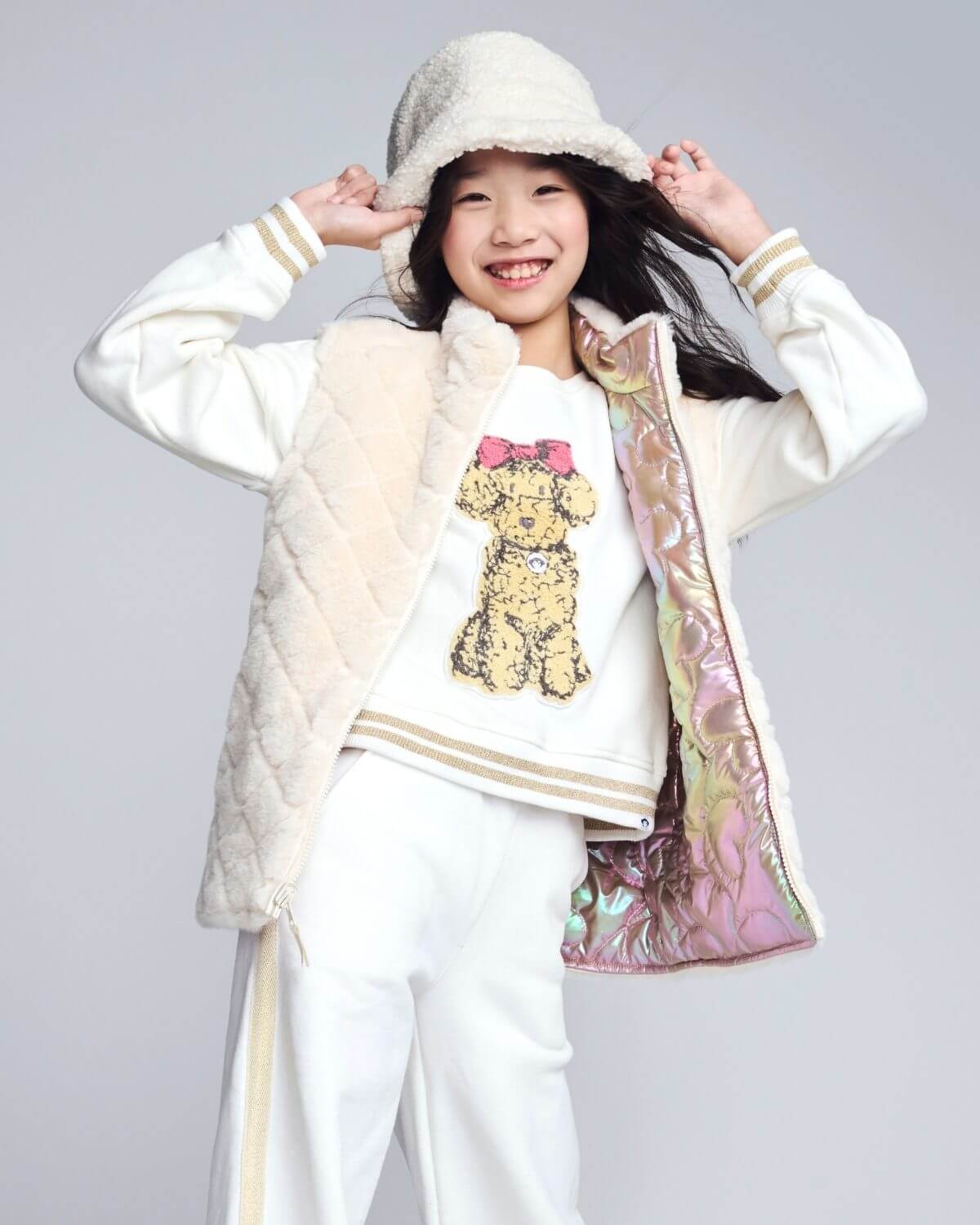 Person wearing a white coat with colorful lining, white pants, and a white shirt with a bear design on a gray background.