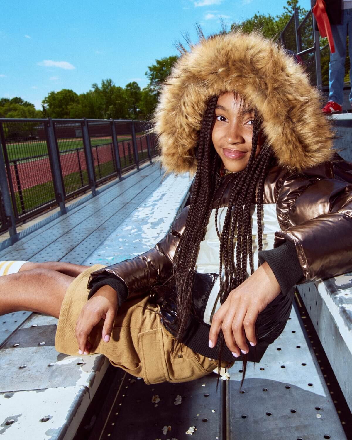 Person wearing a fur-lined coat sitting on a metal structure with a sports field in the background.