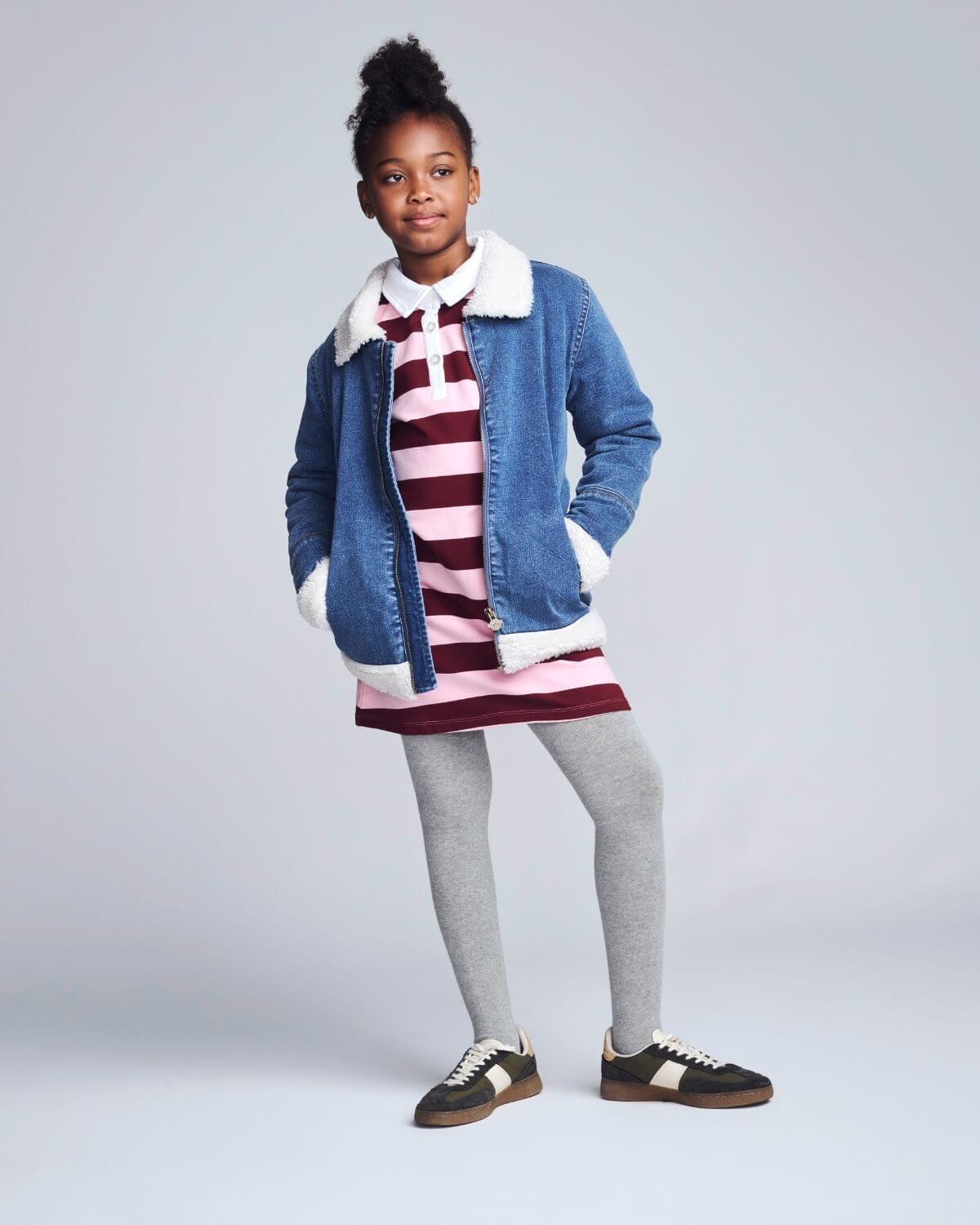 Child wearing a denim jacket with a white fur collar, striped dress, and gray tights on a plain background