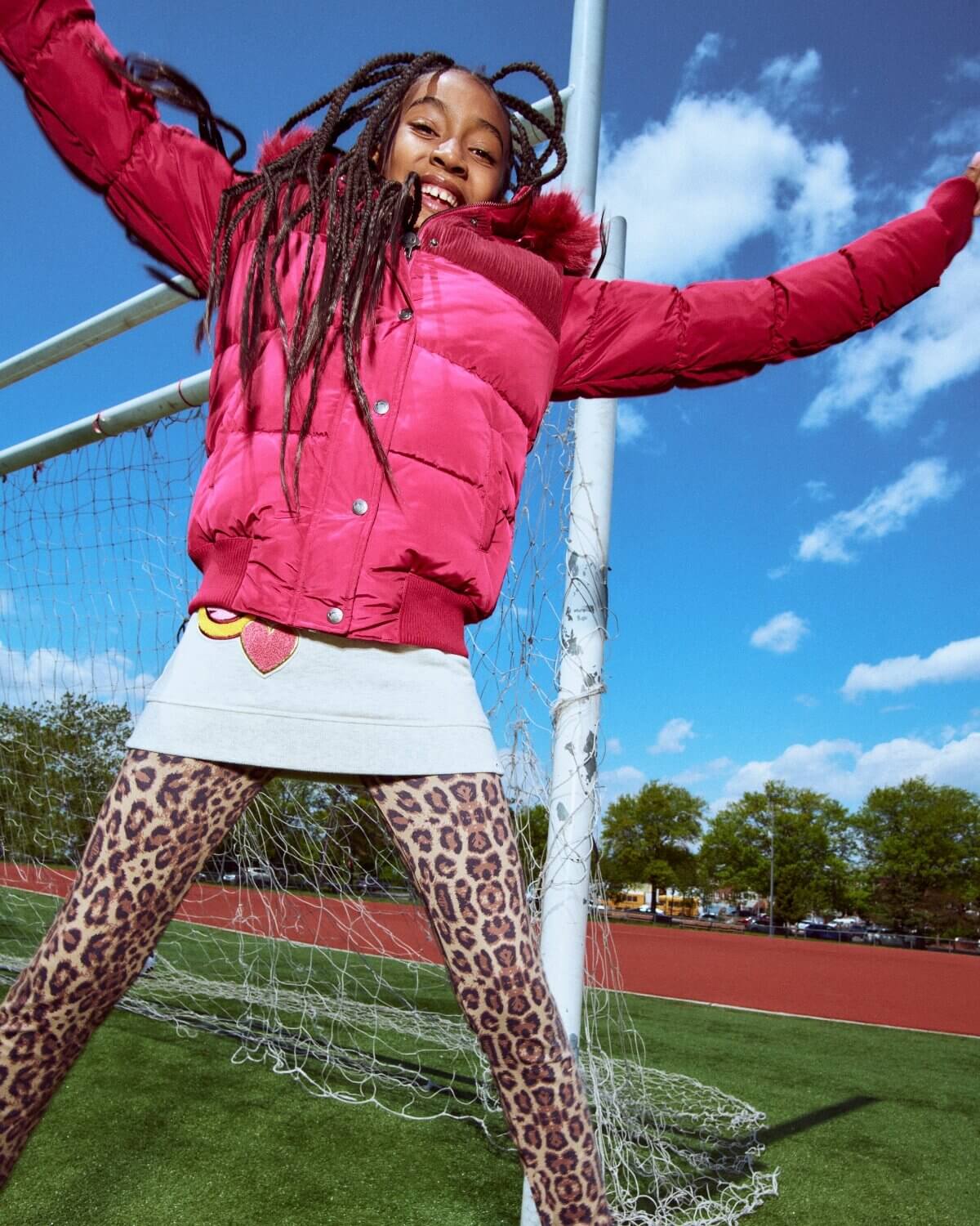 Child in a pink jacket and leopard print pants standing in front of a soccer goal on a sunny day.