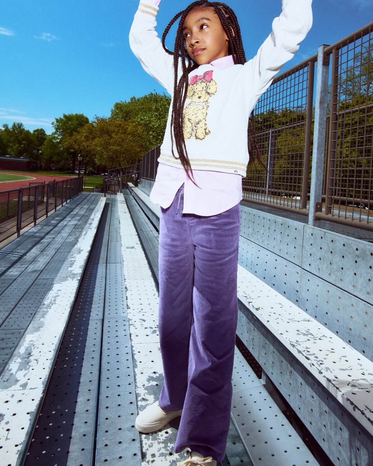 Child standing on bleachers with arms raised, wearing a white sweater and purple pants, against a blue sky.