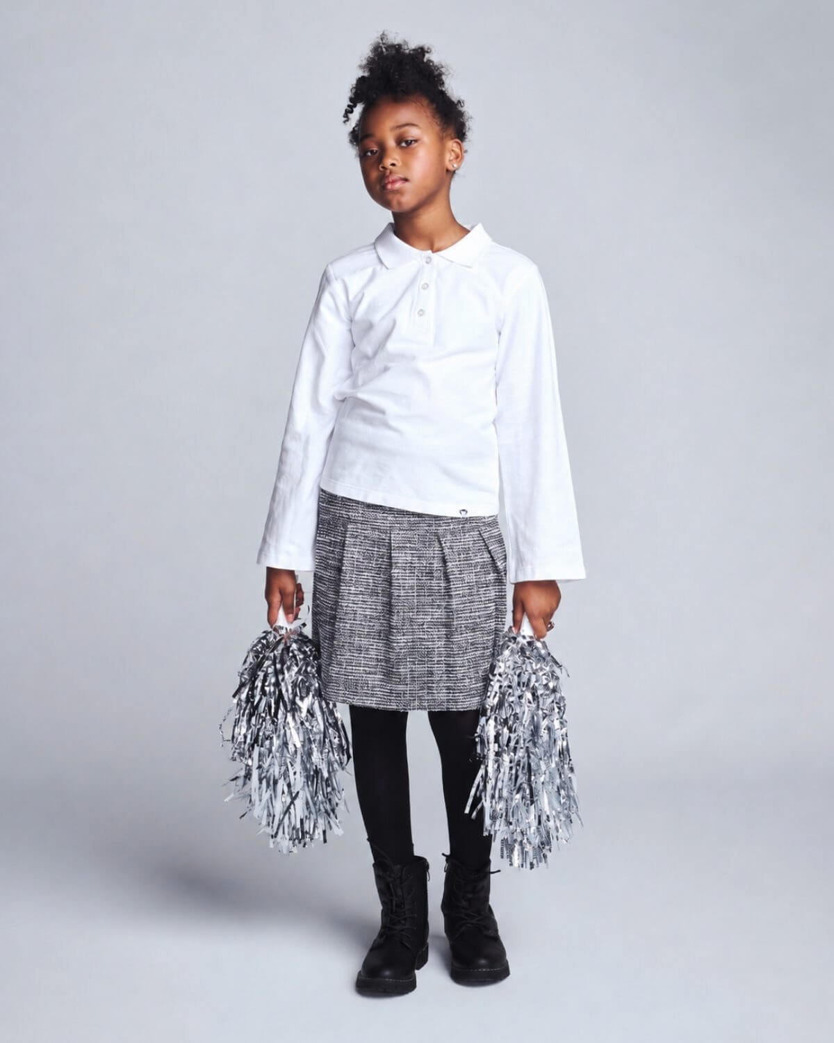 Child wearing a white shirt and gray skirt with pom-poms on a plain background