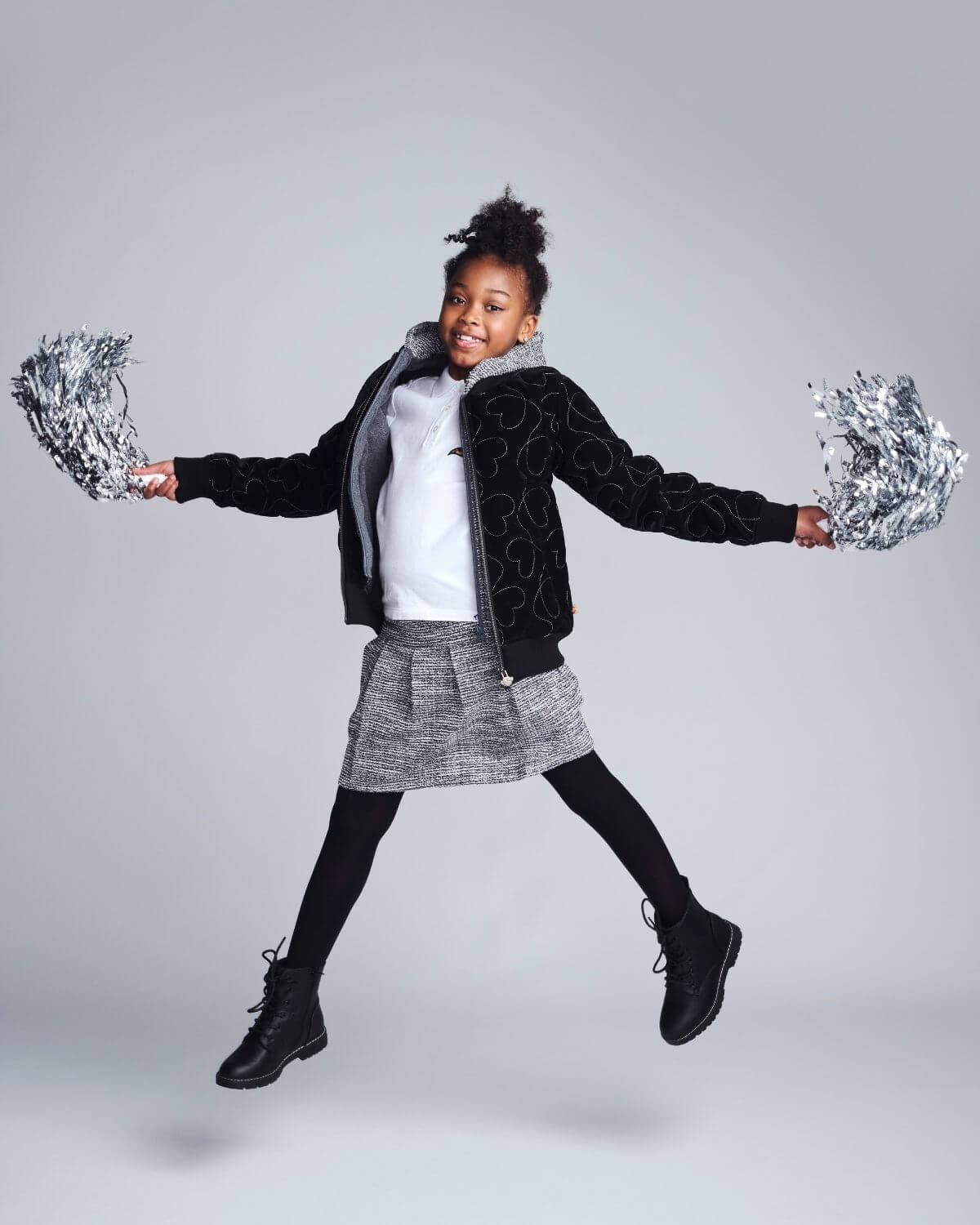 Child in black jacket and gray skirt holding silver pom-poms against a gray background