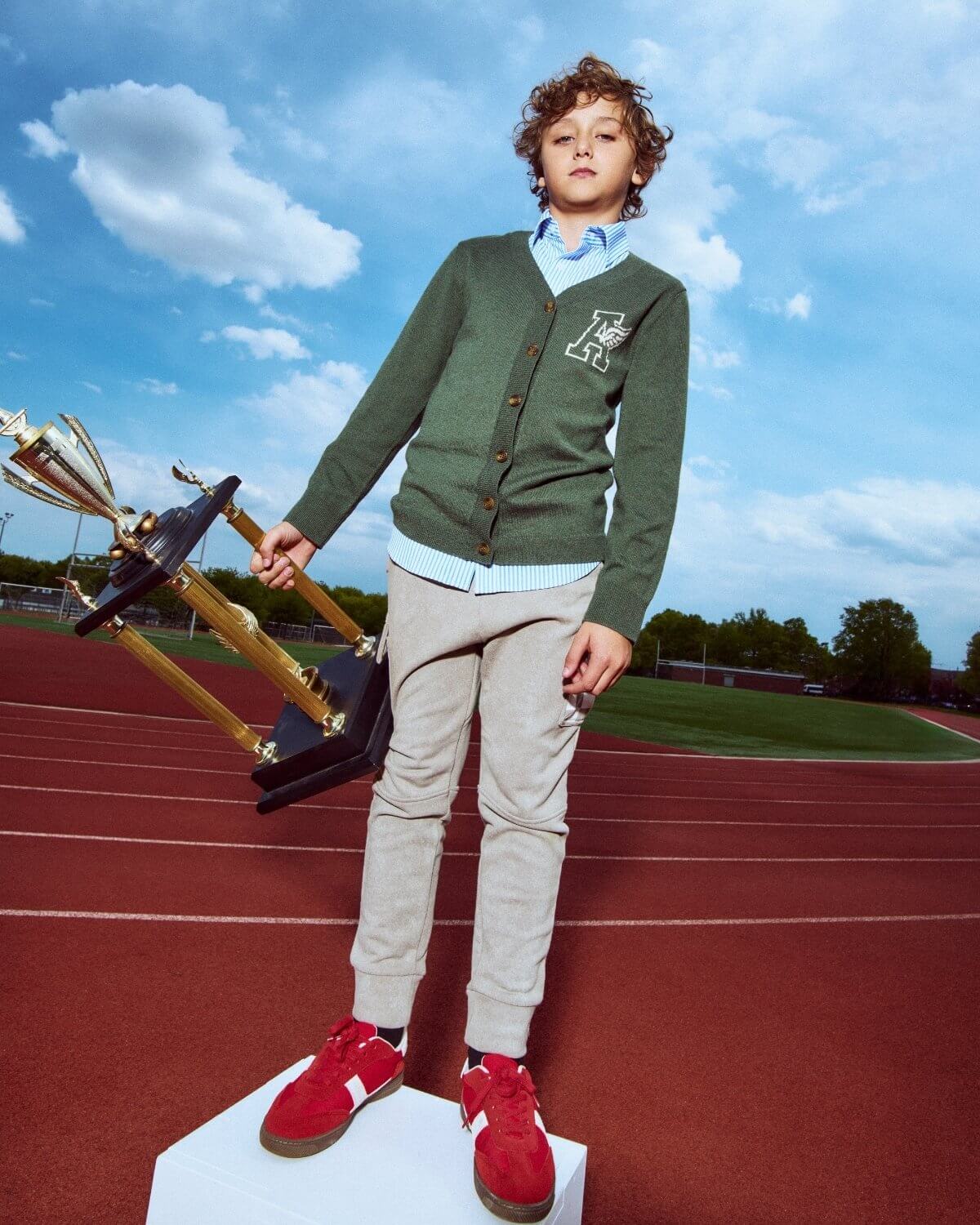 Child in green sweater and red shoes standing on a track with a blue sky background