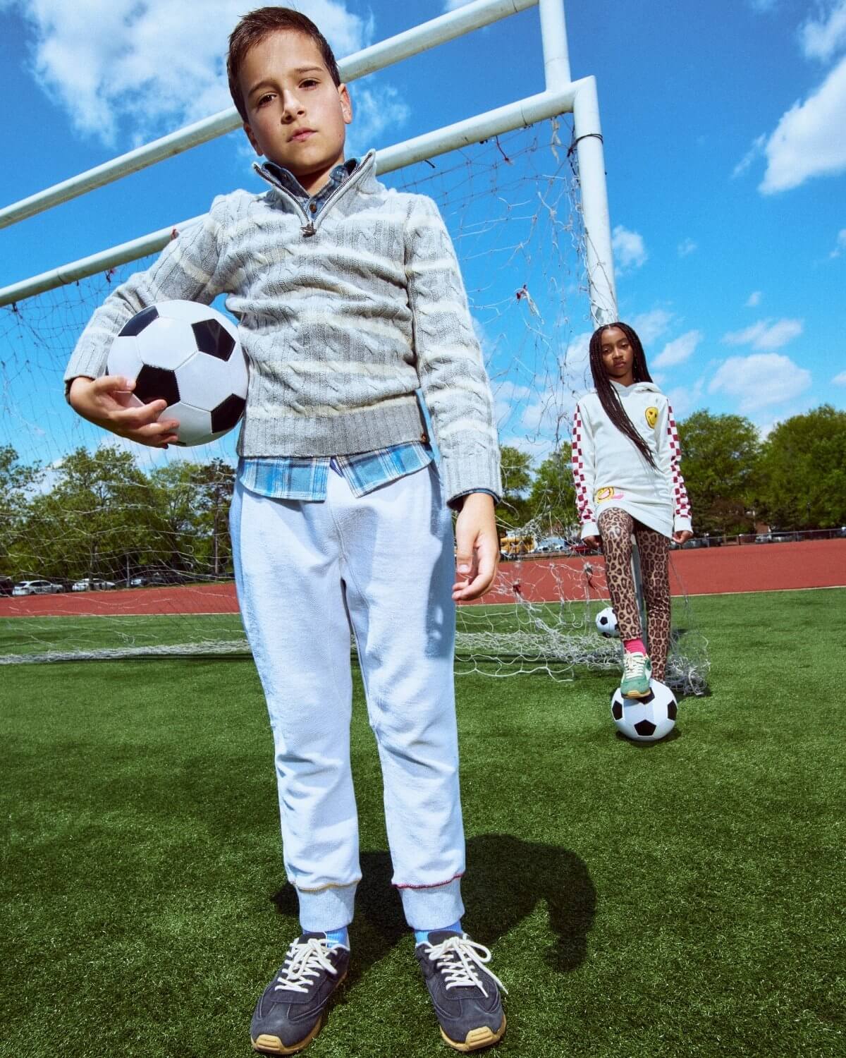 Two children on a soccer field with soccer balls, one holding a ball and the other standing in the background.