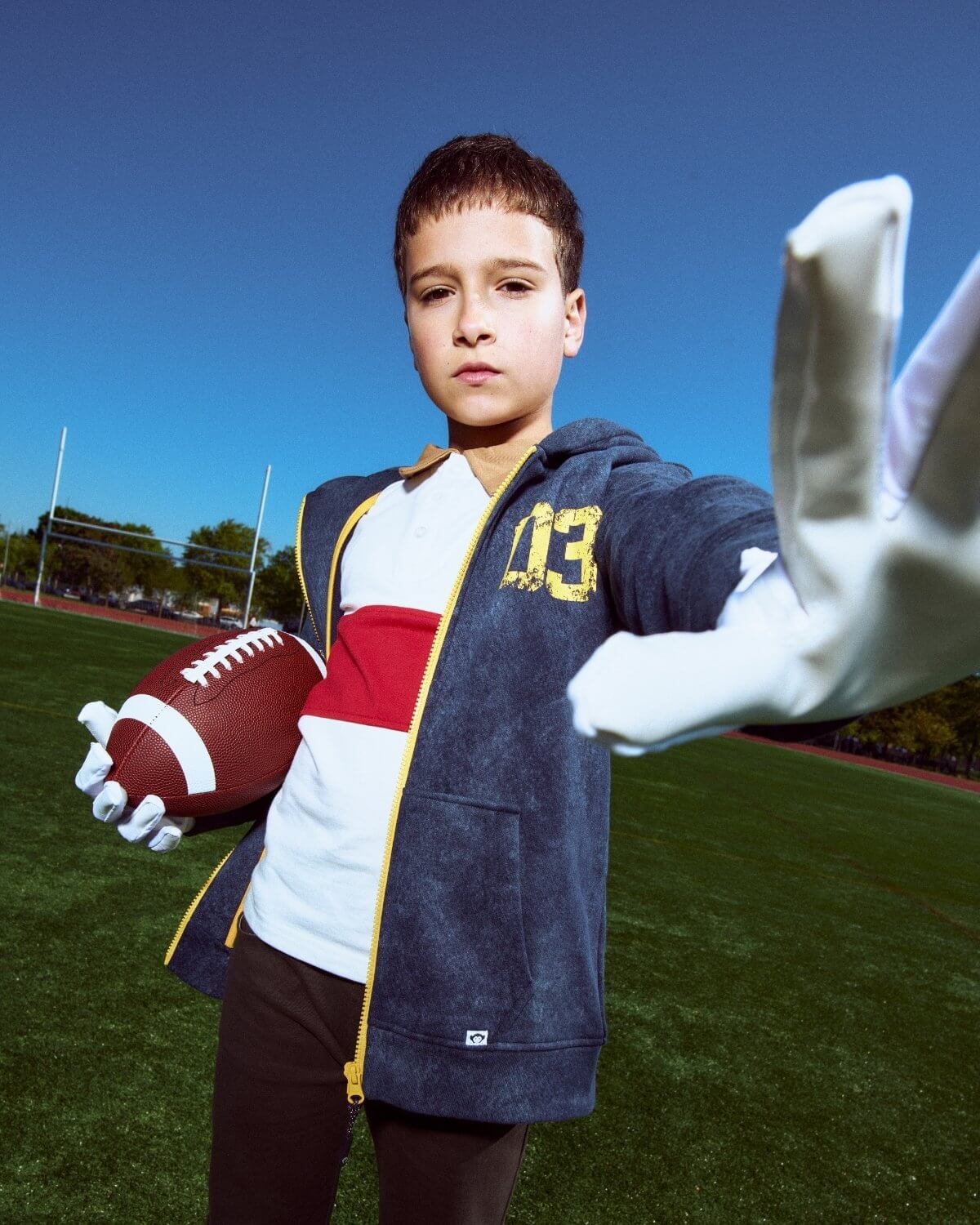 Young boy holding a football and wearing a sports jacket on a sports field.