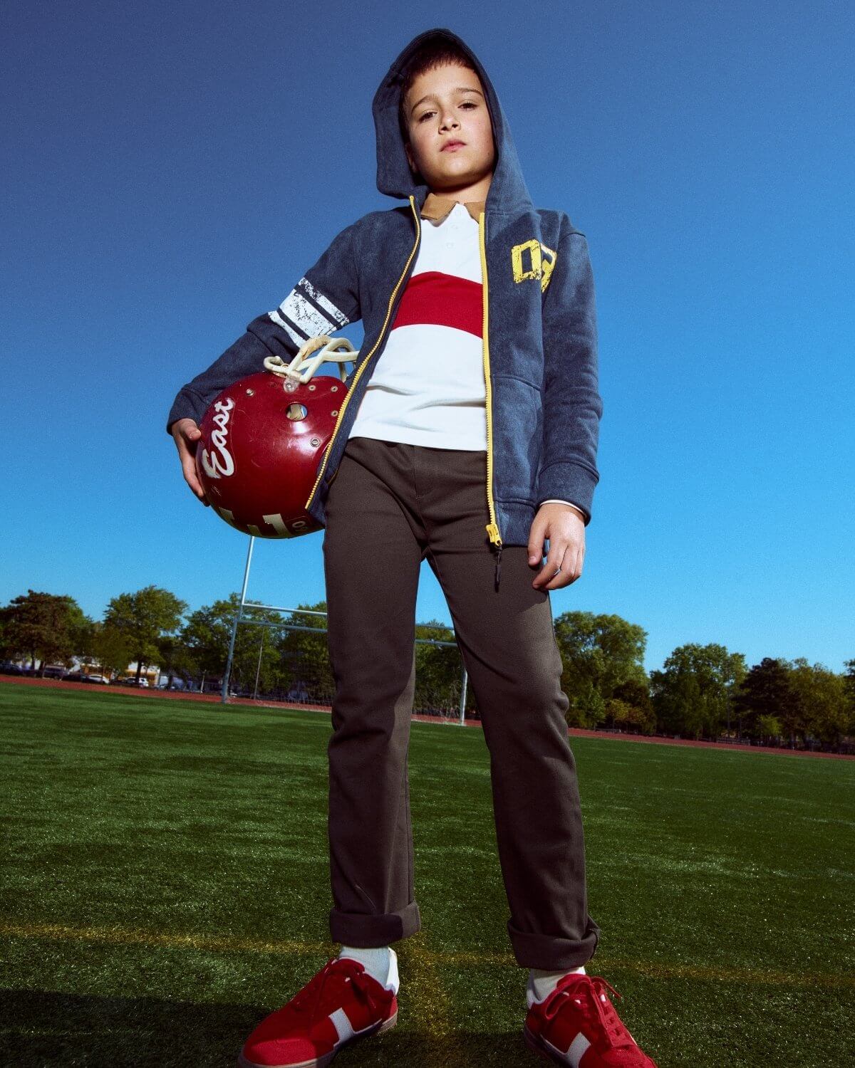 Child holding a football helmet on a sports field with a clear blue sky.
