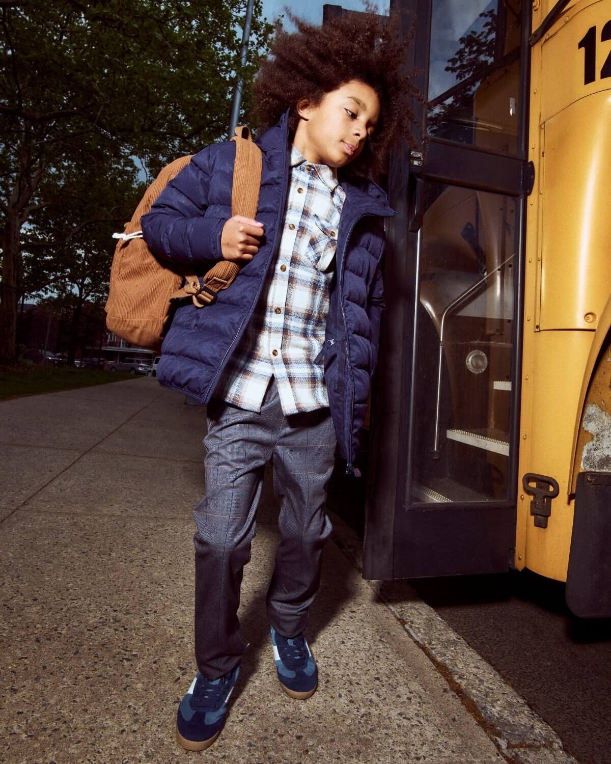 Child getting on a yellow school bus wearing a blue puffer jacket and plaid shirt.