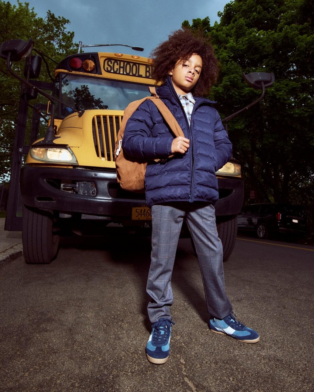 Child standing in front of a school bus wearing a blue jacket and gray pants.