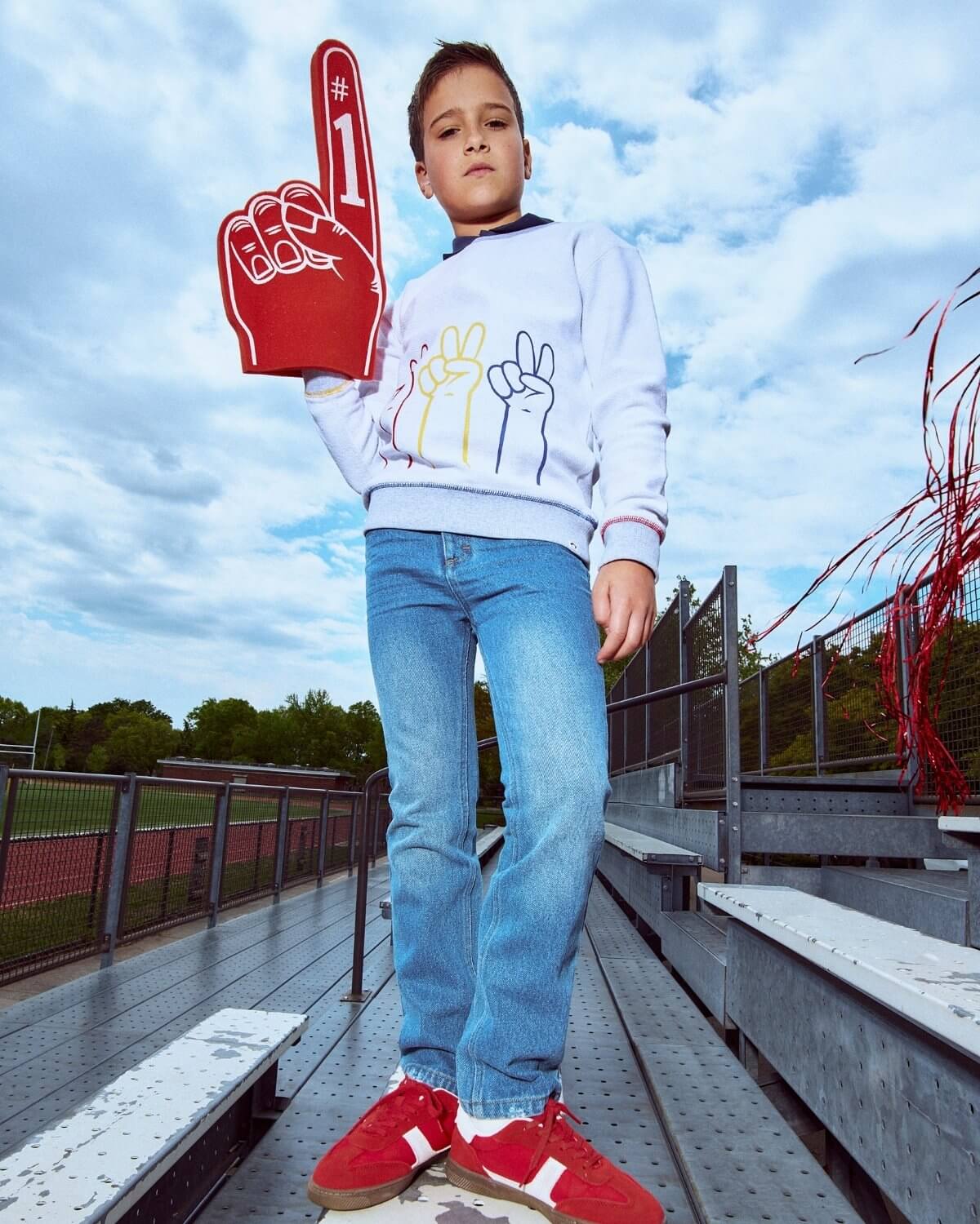 Child holding a foam finger with number 1, wearing a white sweatshirt and blue jeans, standing on bleachers.