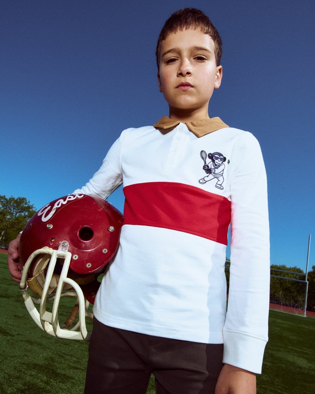 Young boy holding a football helmet on a sports field with a clear blue sky.