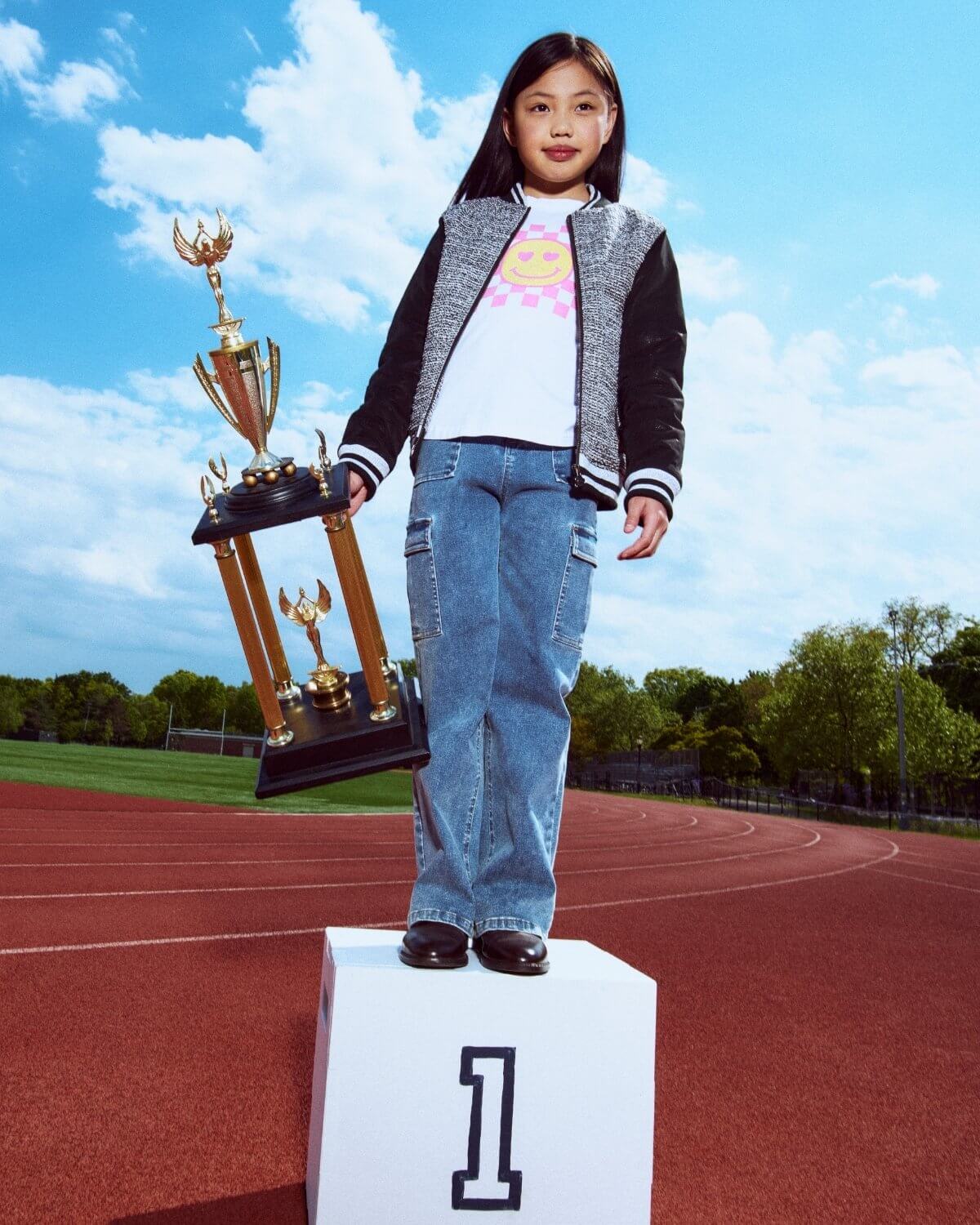 Child standing on a podium with trophies, outdoors on a track and field