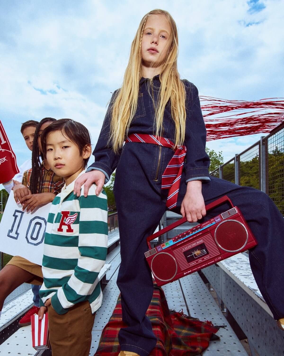 Two children in stylish outfits with a red boombox, standing outdoors against a blue sky.