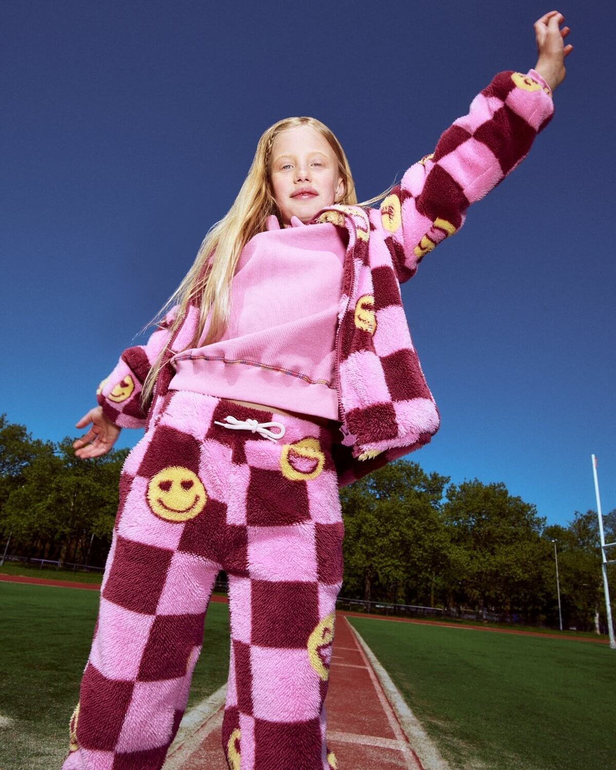 Child in pink and maroon checkered outfit with smiley face designs on a sports field.