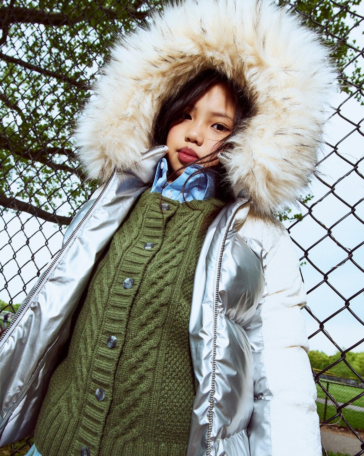Child wearing a silver puffer jacket with a fur-lined hood and a green knitted sweater.