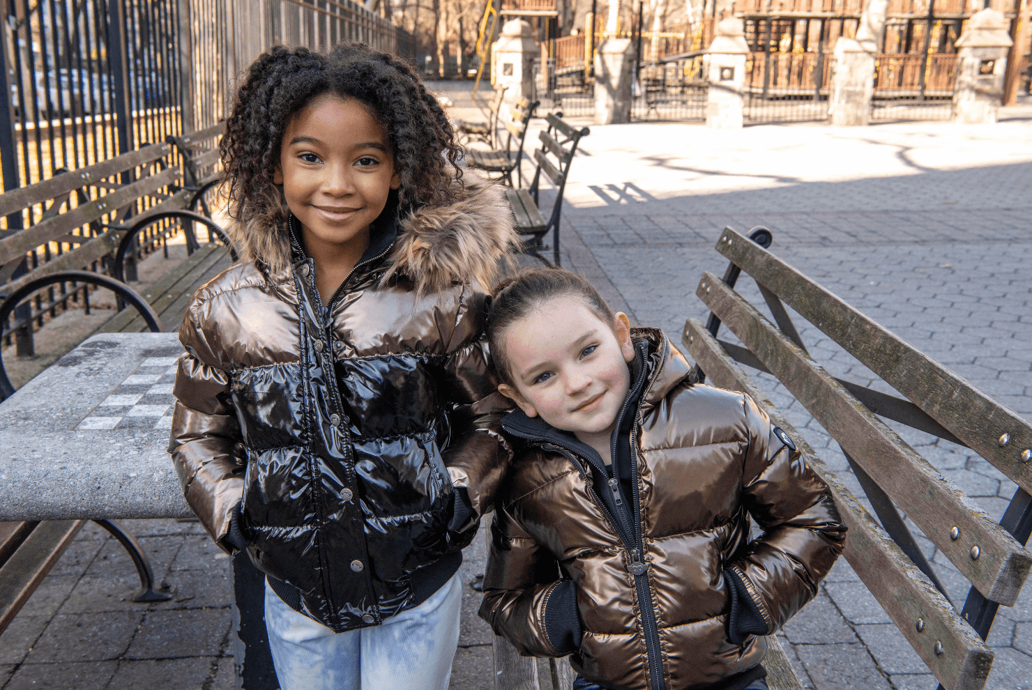 Two stylish kids in shiny puffy jackets enjoying a sunny day in the park, showcasing playful winter fashion.