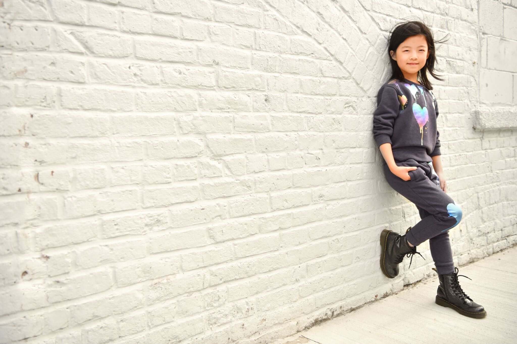 Stylish girl in playful Appaman outfit, showcasing trendy kids' fashion against a chic white brick wall.