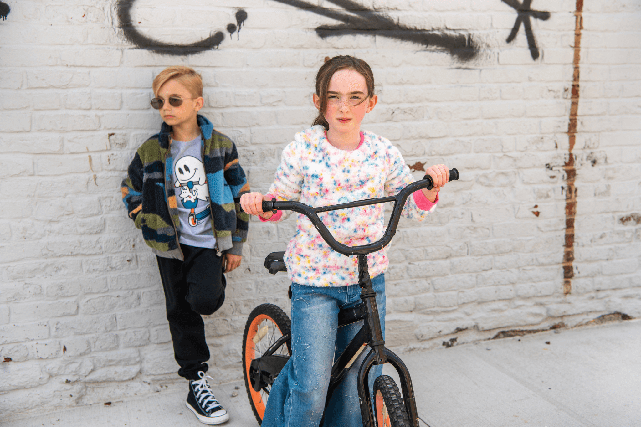 Two stylish kids, one on a bike and the other with sunglasses, showcasing trendy back-to-school outfits in a graffiti setting.