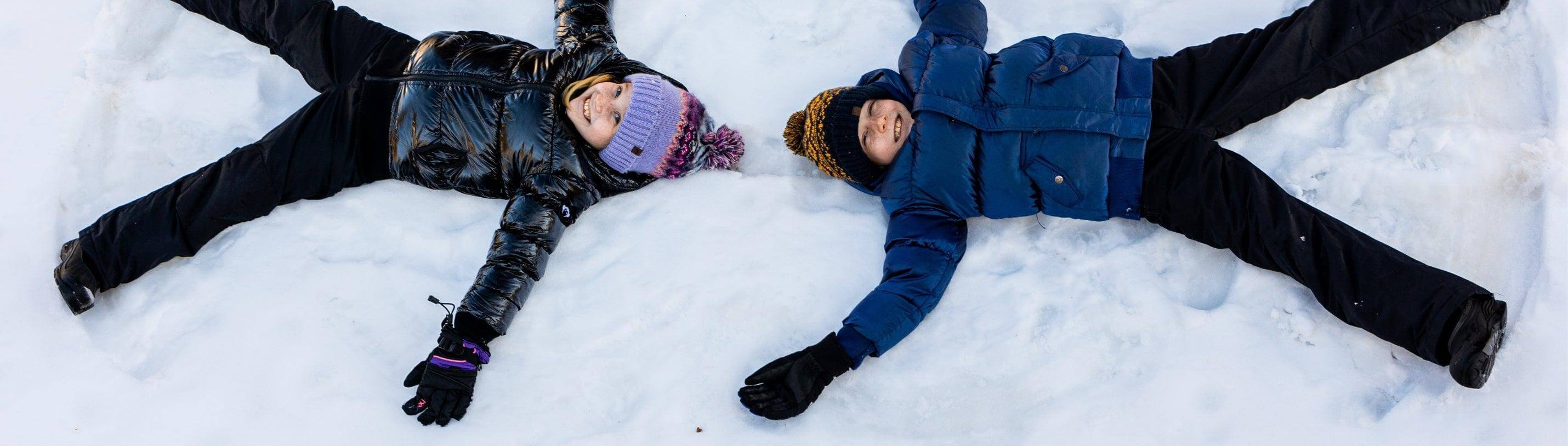 Two kids joyfully making snow angels in a winter wonderland, bundled up in stylish jackets and gloves, embracing the season's fun.