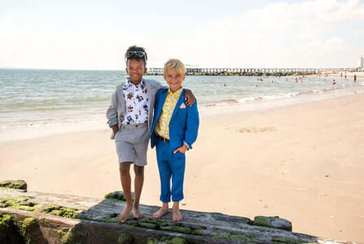 Two stylish kids in vibrant suits enjoying a beach day, showcasing playful summer suiting options for boys.
