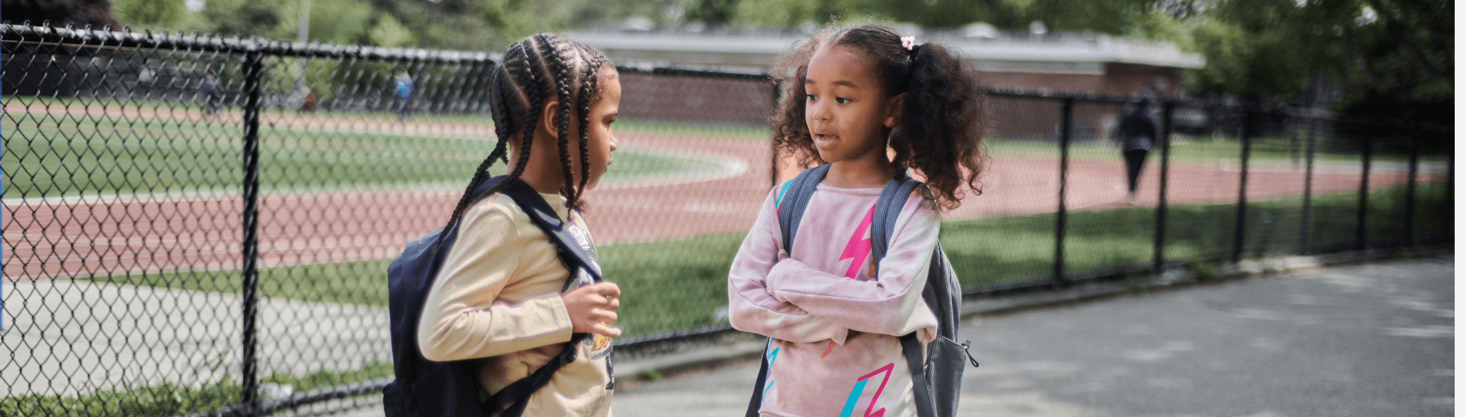 Two stylish girls chatting outdoors, showcasing trendy outfits and backpacks for school.