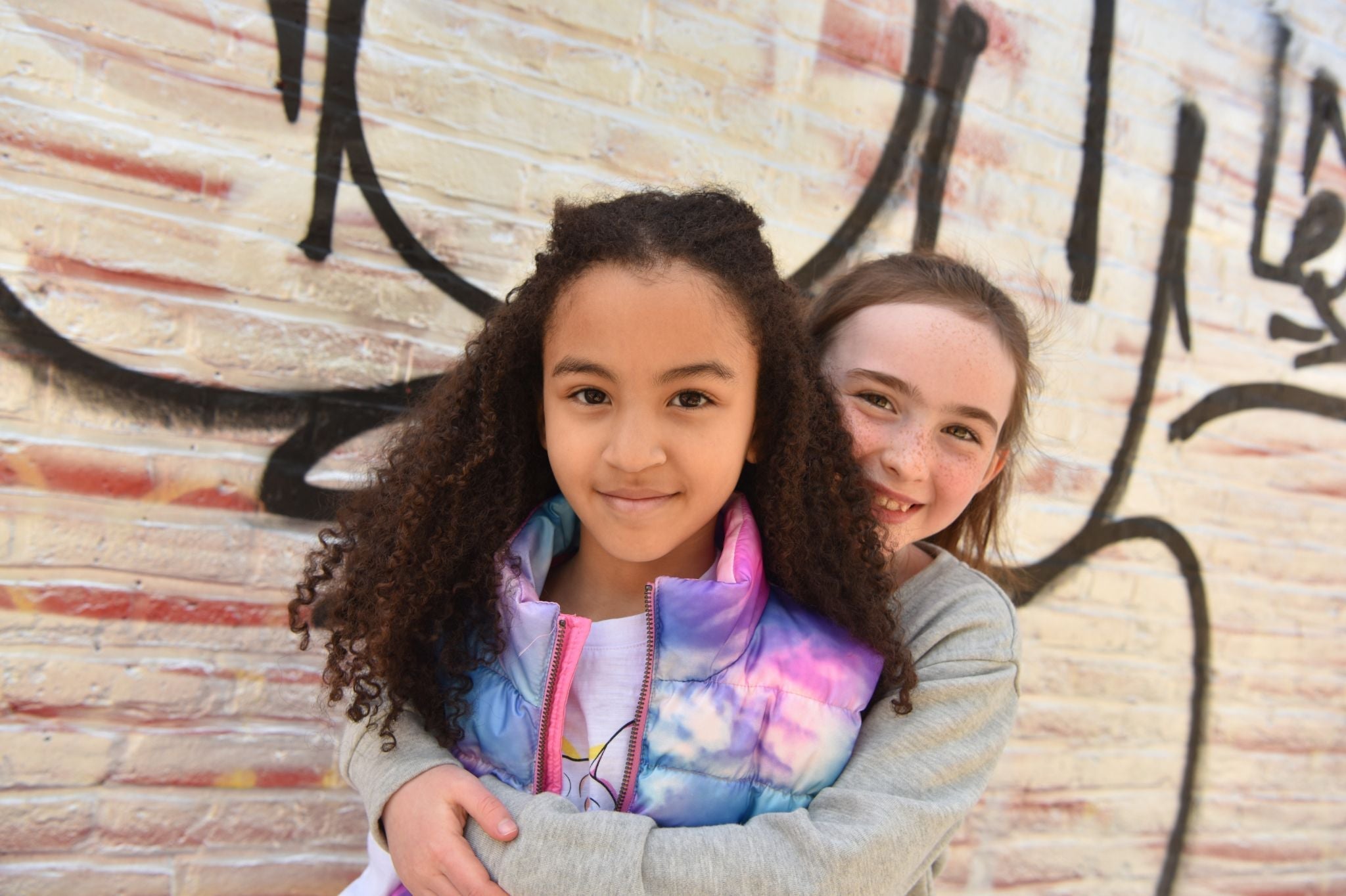 Two stylish girls sharing a joyful moment, rocking colorful Appaman outfits against a vibrant graffiti wall.