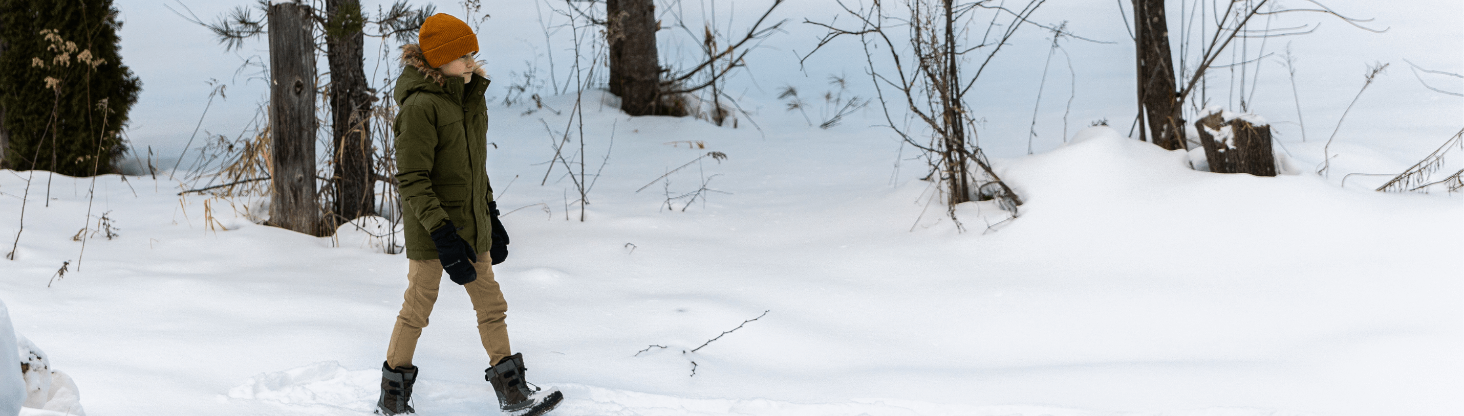 A stylish kid in a cozy green jacket and orange hat walks through snowy woods, ready for family winter fun.