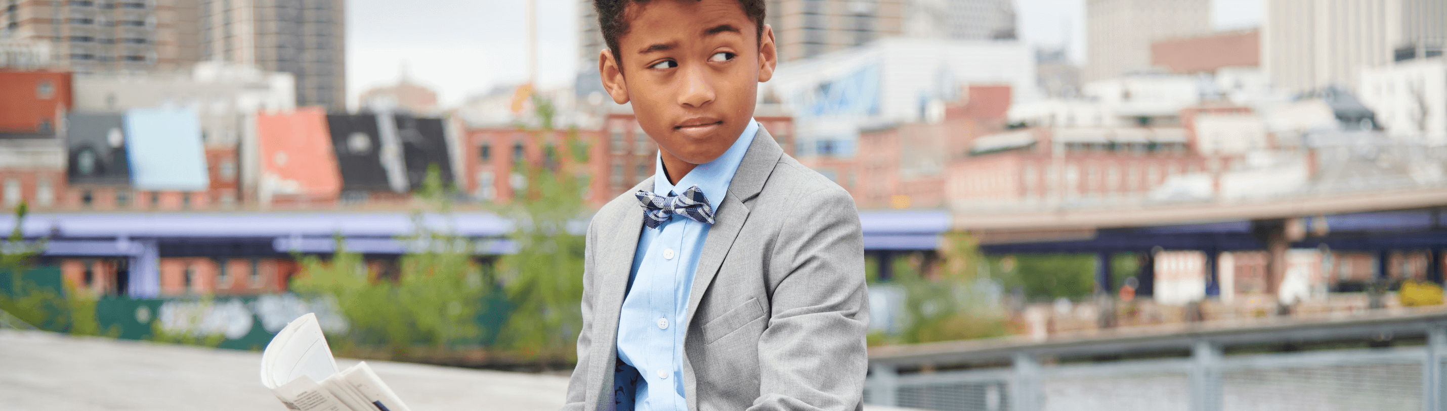 Dapper young boy in a gray suit and bow tie, showcasing stylish kids fashion in a vibrant urban setting.