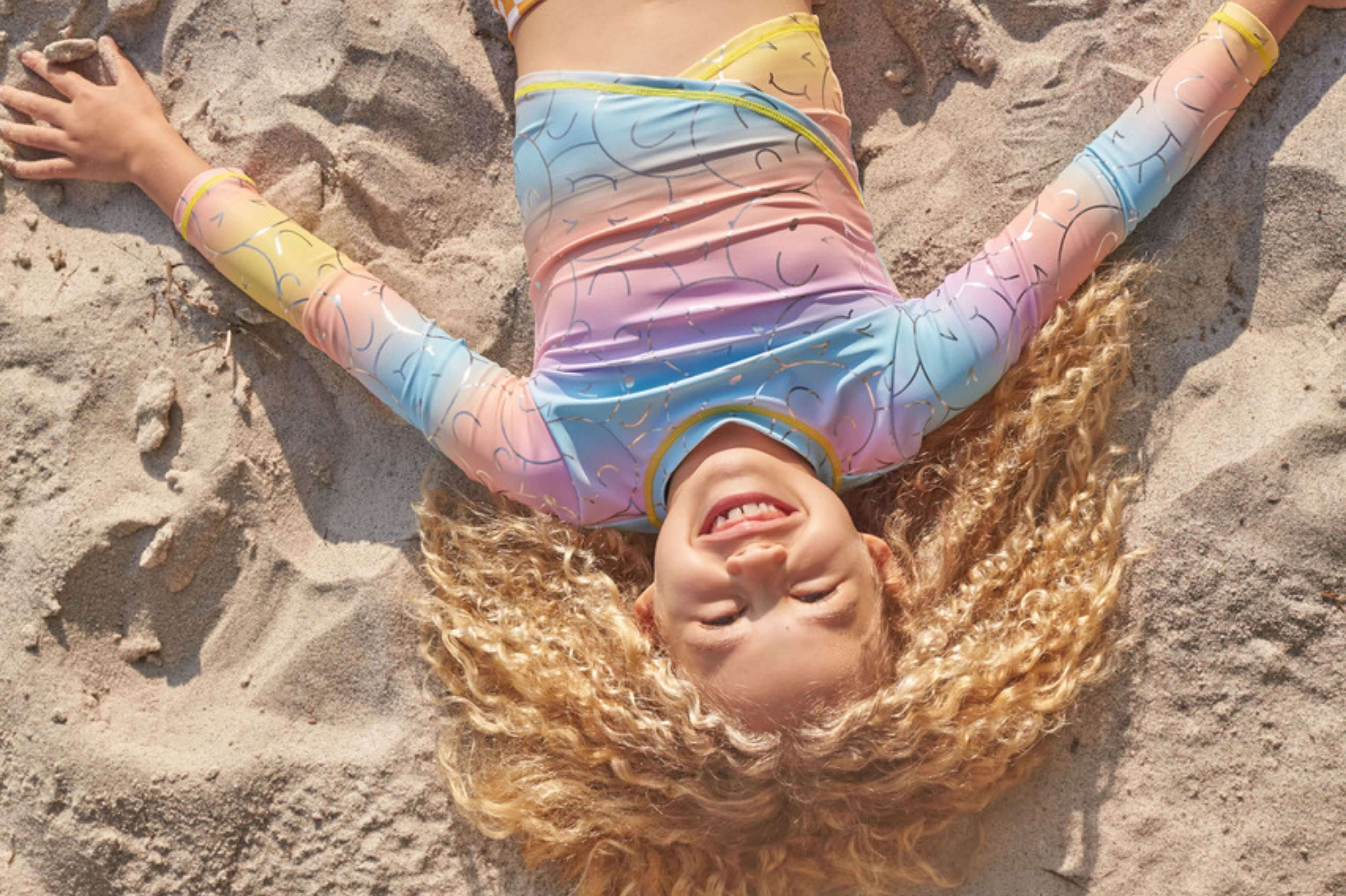 A joyful child in a colorful swimsuit lying on the sand, embodying carefree summer vibes with playful, beach-ready style.
