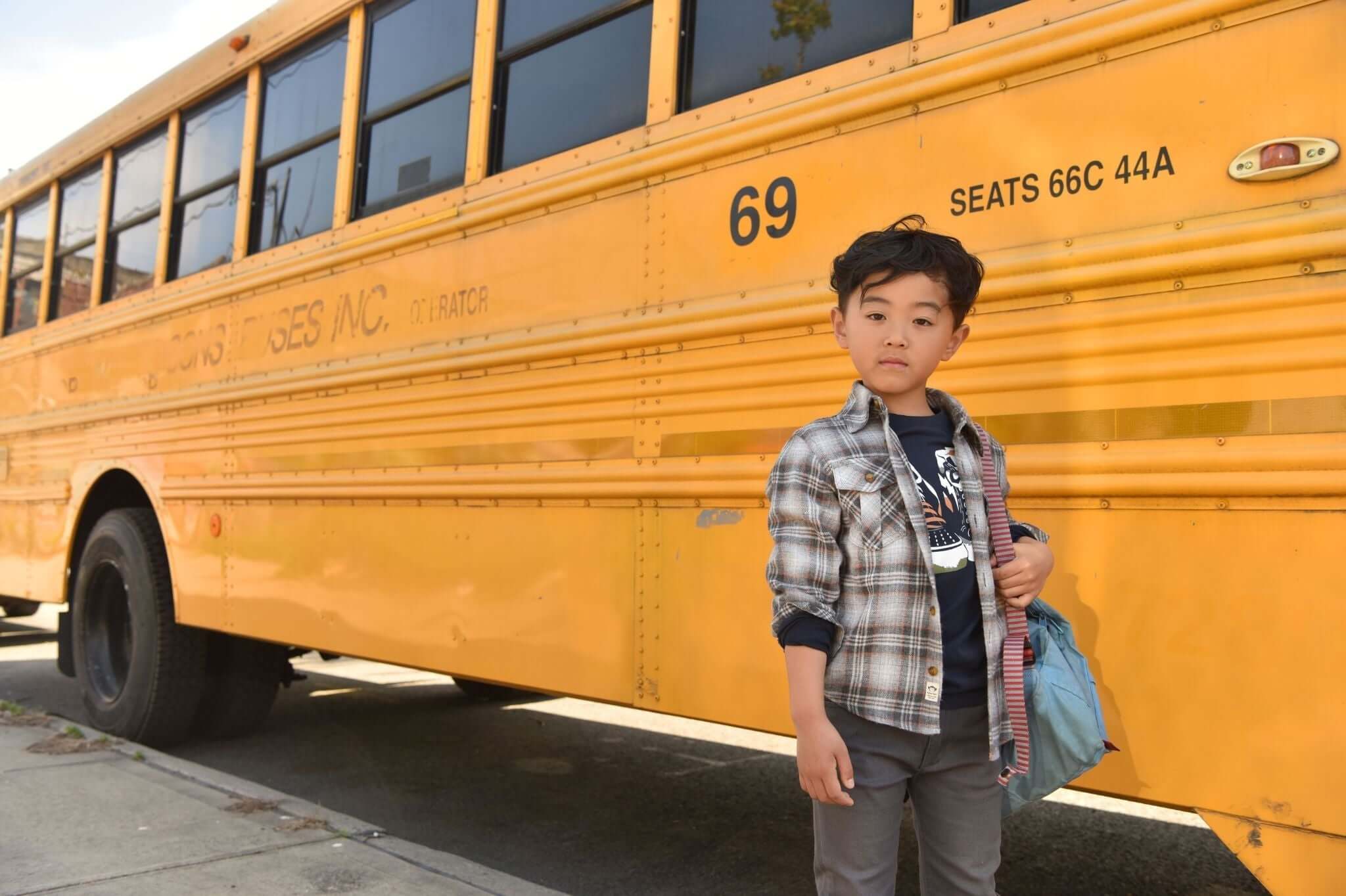 Stylish boy in a plaid jacket stands next to a bright yellow school bus, showcasing trendy kids' outfit ideas.