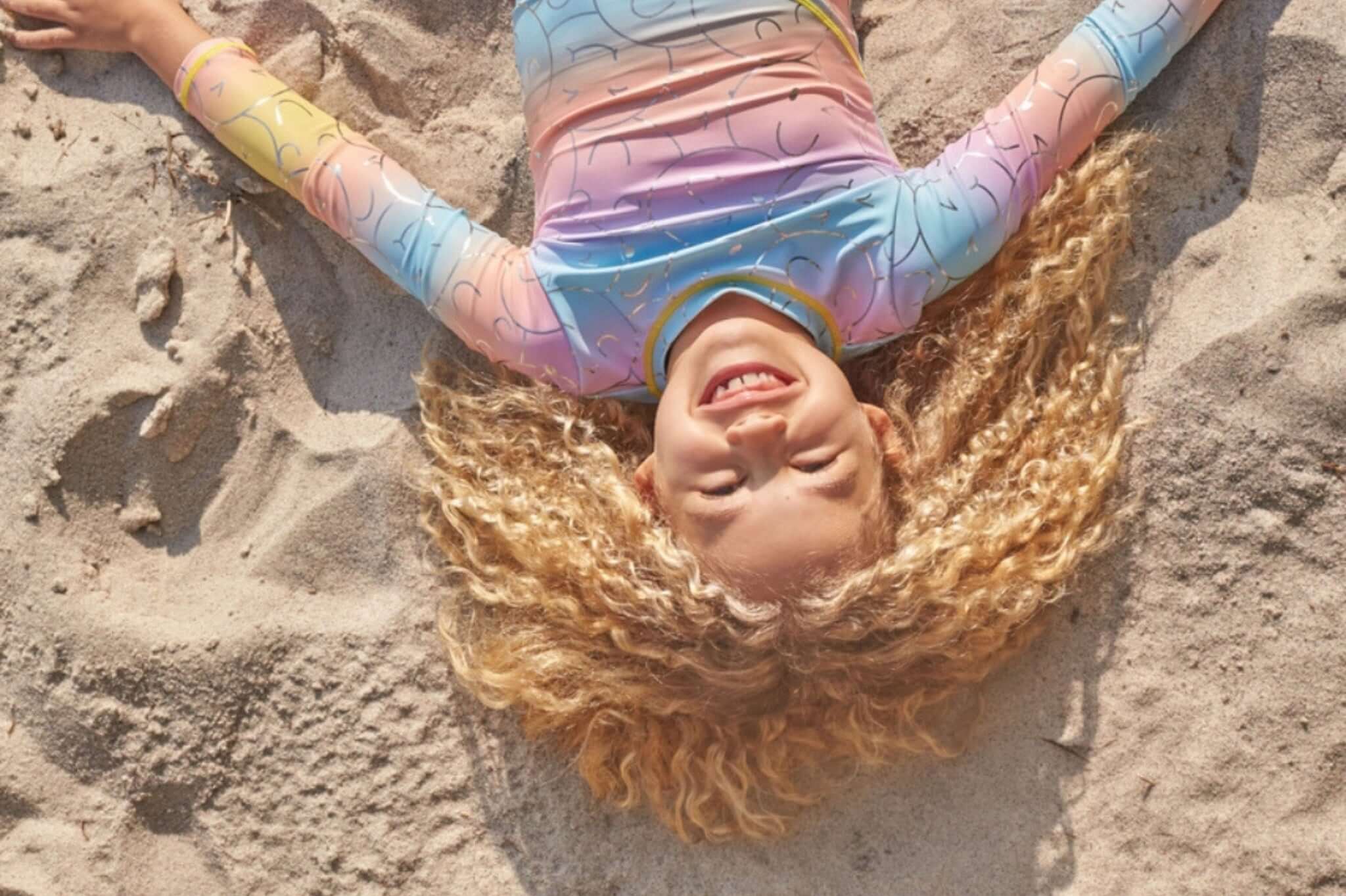 A joyful girl with curly hair smiles widely while lying on the sandy beach, showcasing her colorful swimsuit.