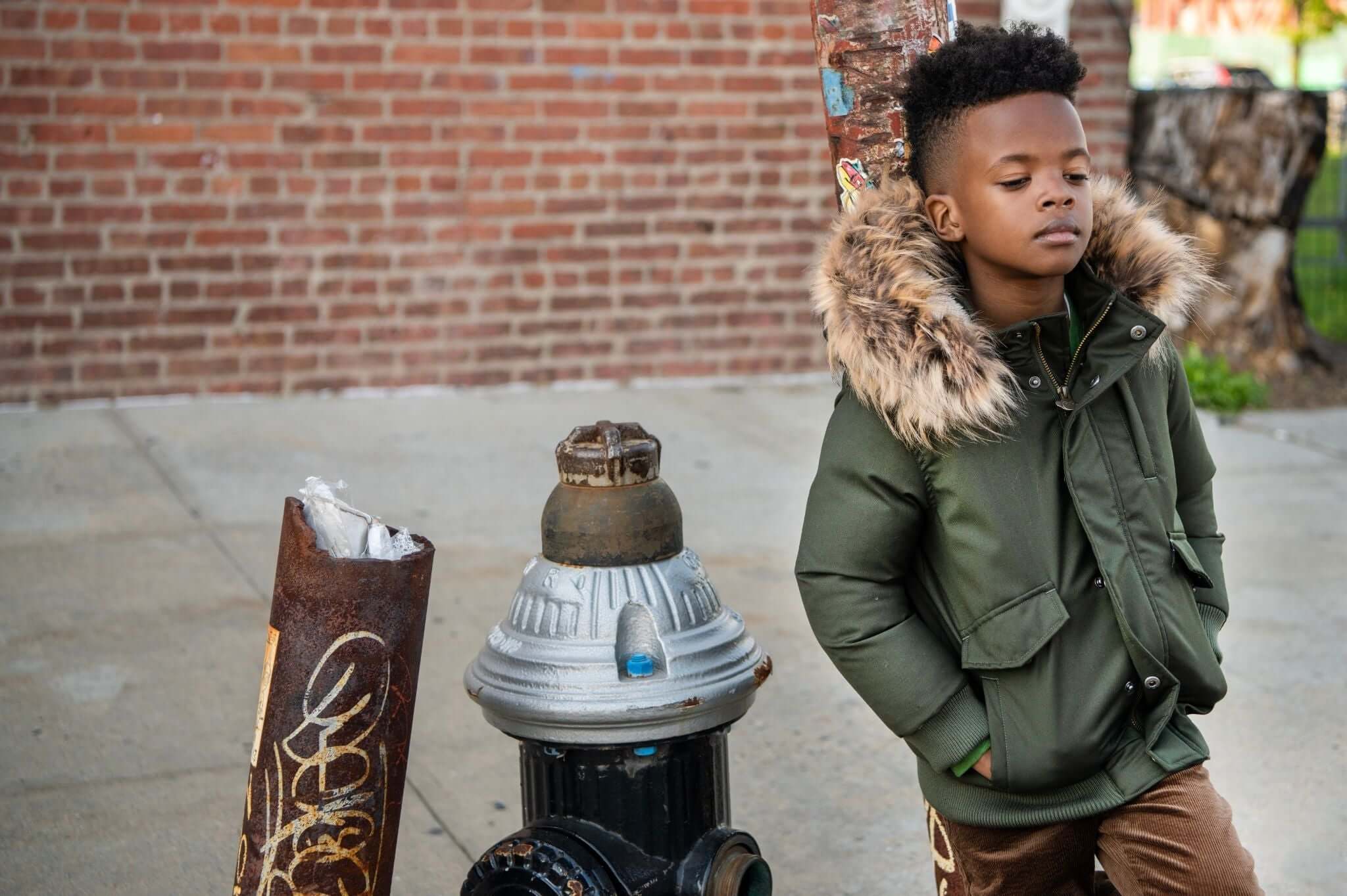 Stylish boy in a green down jacket with fur trim, posing confidently by a graffiti-covered fire hydrant.