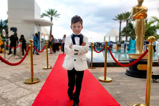 Stylish young boy in a tuxedo walks confidently on the red carpet, ready to steal the spotlight at a glamorous event.