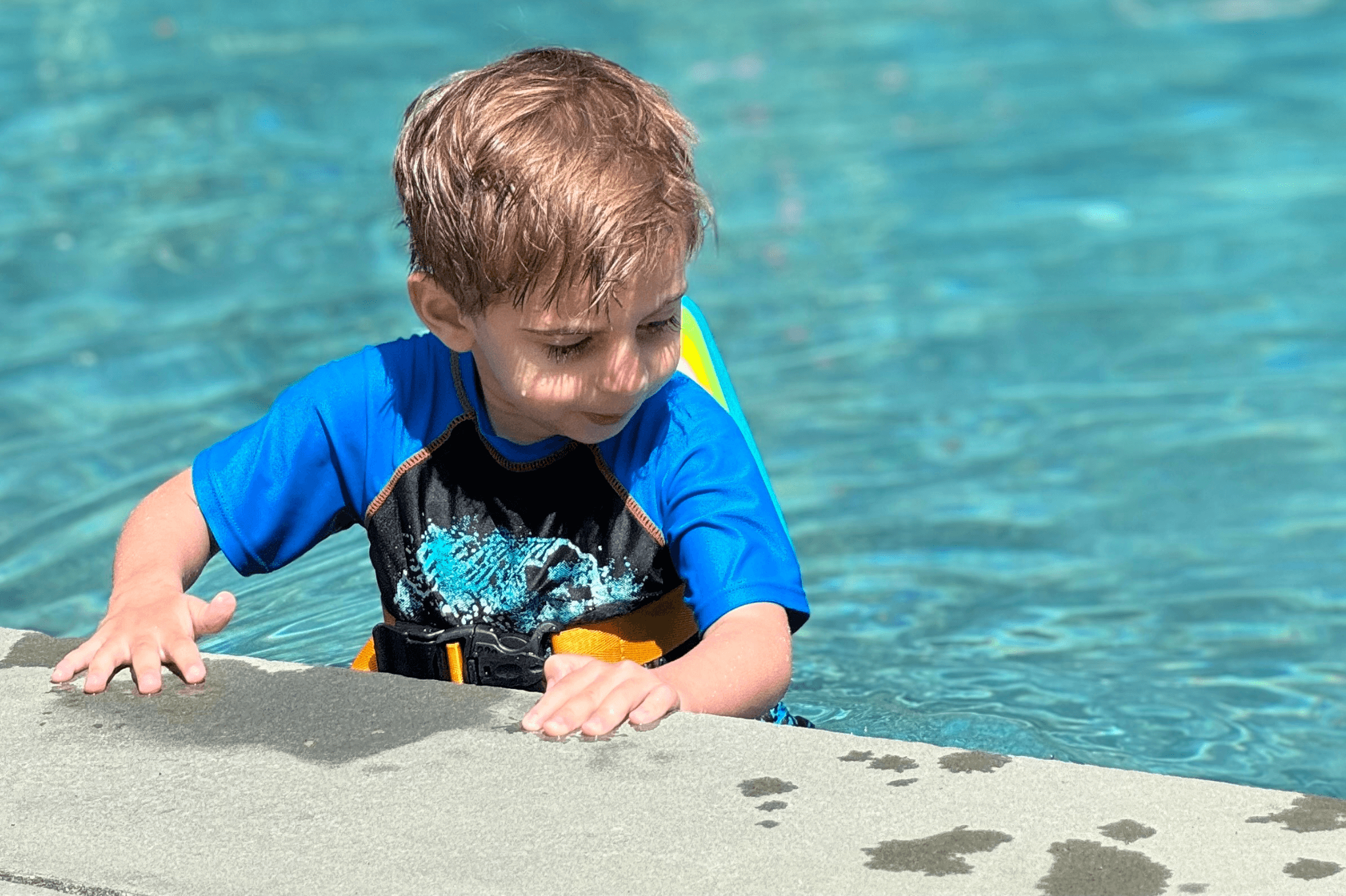 A young boy in a stylish swim shirt joyfully splashes at the edge of a sparkling pool, ready for summer fun.
