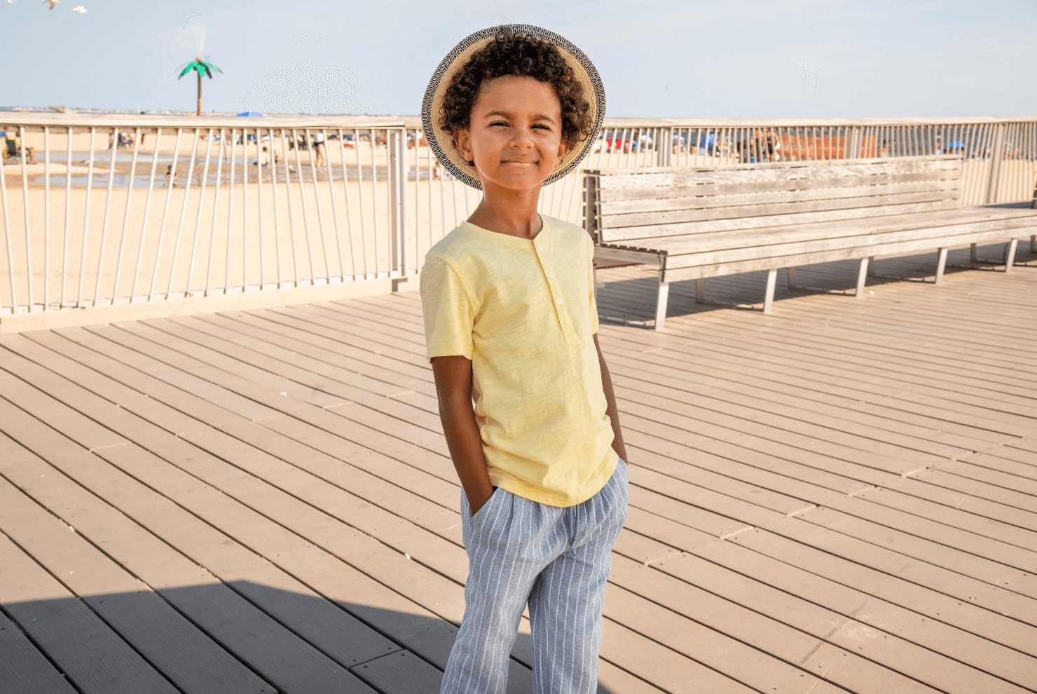 Stylish boy in a sunny yellow tee and striped pants, radiating confidence at the beach—parent-approved summer vibes!