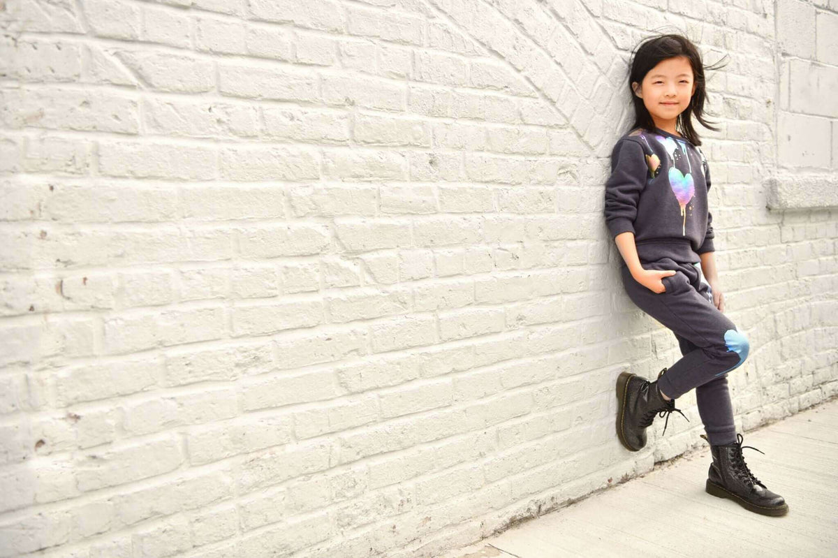 Stylish girl in playful Appaman outfit, showcasing trendy kids' fashion against a chic white brick wall.
