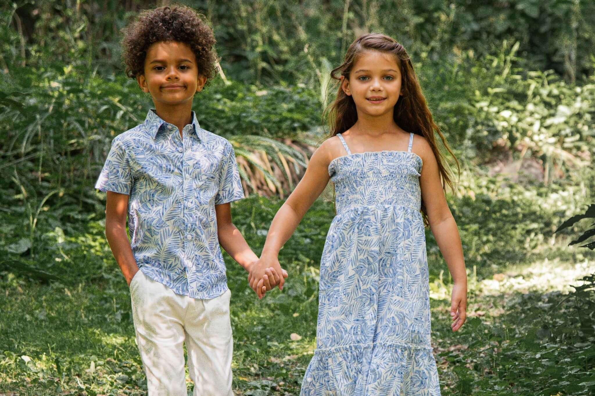 Stylish kids' summer outfits: a boy in a floral print shirt and white pants, and a girl in a blue dress, holding hands in nature.