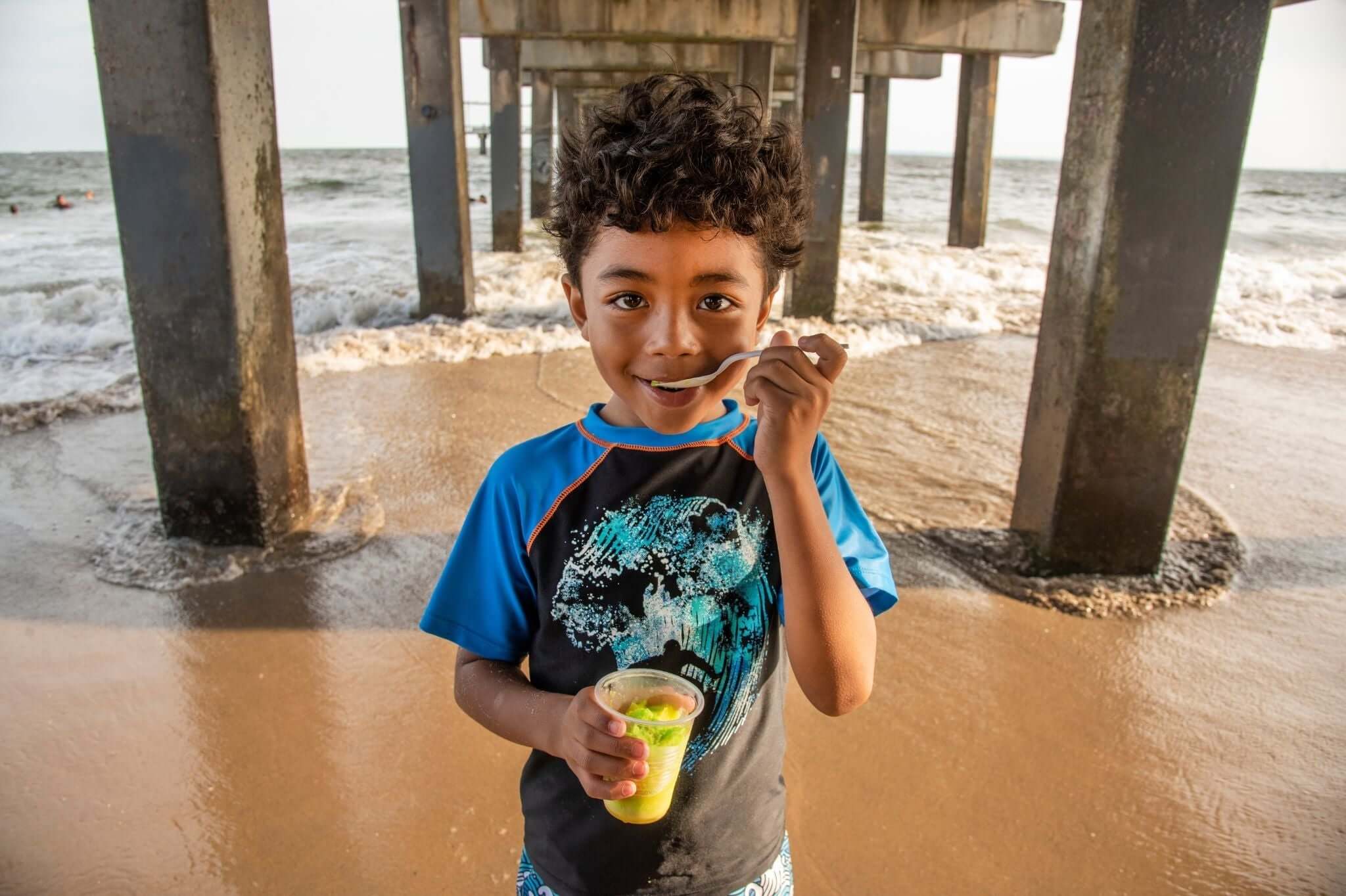 Smiling boy enjoying a refreshing treat under a pier, wearing colorful swimwear, perfect for summer family fun!