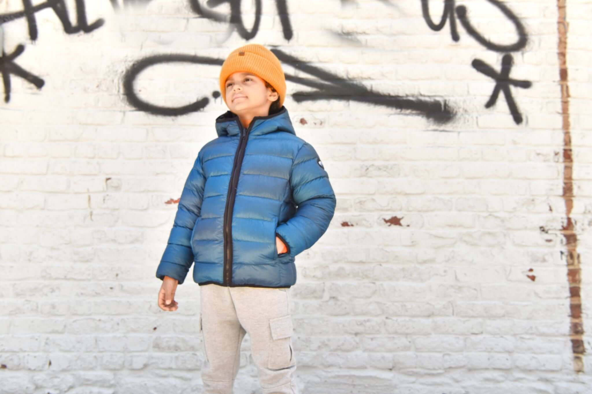 Stylish boy in a blue puffer jacket and orange beanie, showcasing trendy kids' winter fashion against a graffiti wall.