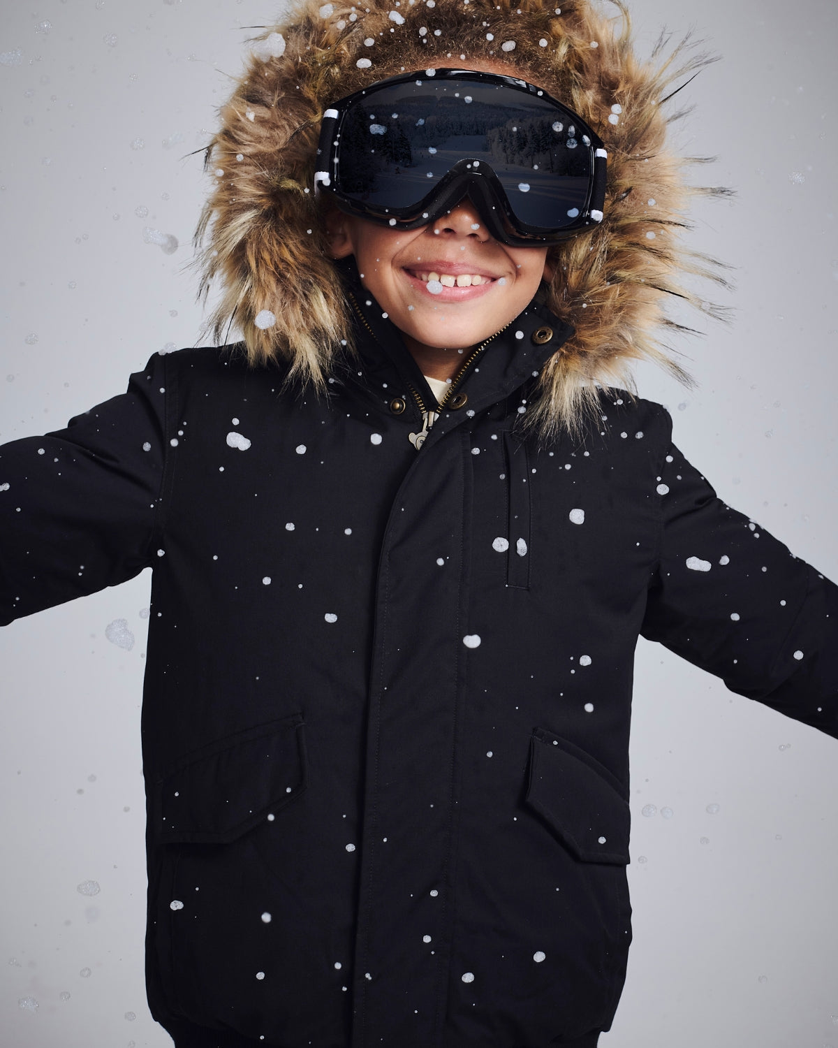 Happy child wearing a black Kids' Down Coat with fur hood and ski goggles in falling snow.