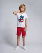 Child wearing a white t-shirt with a colorful graphic and red shorts on a gray background