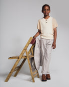 A child wearing a Beige Sweater Knit Polo with collared neckline, paired with light gray pants, standing by a wooden chair.